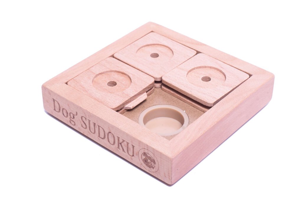 Sur la photo, un petit chien (ou un chat) est en train de jouer avec le PET' SUDOKU SMALL BASIC. Le jouet, un puzzle en bois avec plusieurs compartiments coulissants, est posé au sol. L'animal, concentré et curieux, utilise sa patte pour déplacer l'un des couvercles du puzzle dans le but de découvrir une friandise cachée à l'intérieur. Le fond de l'image est un intérieur chaleureux avec un sol en bois clair, accentuant la sensation de confort et de détente.